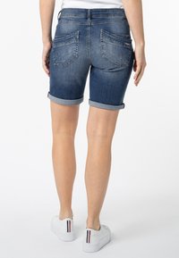Denim bermuda shorts in een donkere wassing met opgerolde boorden, voorzien van twee achterzakken en subtiele vervaging. Gecombineerd met witte sneakers.