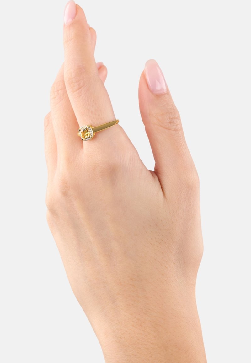 Anillo de oro amarillo con una piedra amarilla de corte cuadrado, rodeada de pequeñas piedras transparentes. Banda lisa, llevado en una mano parcialmente visible.
