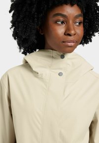 Beige vattentålig jacka med hög krage, utrustad med två tryckknappar och en ren, minimalistisk design, lämplig för olika väderförhållanden.