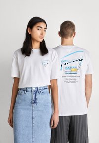 Camiseta de algodón blanca con un diseño de bolsillo en el frente y estampados coloridos en la parte posterior. Modelo combinado con una falda de mezclilla y pantalones a rayas.
