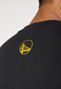 Μαύρο βαμβακερό μπλουζάκι με κίτρινο λογότυπο γέφυρας στο πάνω μέρος της πλάτης. Απλή σχεδίαση με στρογγυλή λαιμόκοψη και λεία υφή.