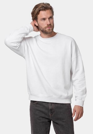 Skægget mand med lyst brunt hår iført en plain hvid sweatshirt og mørke jeans, der poser med den ene hånd bag hovedet.