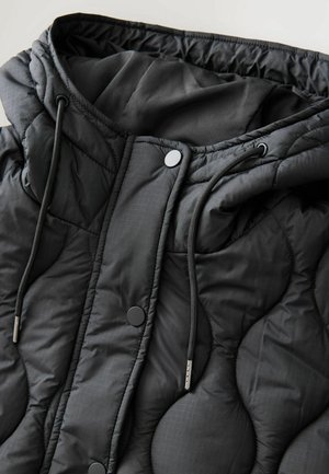 Chaqueta acolchada negra con capucha, con una textura suave, cierre con cordón y botones a presión. Patrón de cuadrícula sutil.