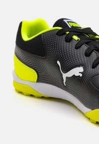 Sorte og grå syntetiske sportsko med et bølget mønster, neongule detaljer og et hvidt Puma-logo. Har en struktureret overdel og gummisål.
