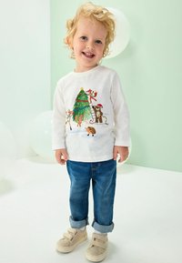 Chemise blanche à manches longues avec un imprimé coloré d'animaux et d'arbres, associée à un jean en denim bleu et des baskets beiges à velcro. Fond clair.