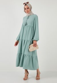 Light teal, long-sleeve dress featuring tiered design, tassel detail, and soft texture. Paired with a beige clutch and heels.