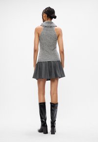 Donna con capelli intrecciati che indossa un top a maniche corte grigio in maglia, una mini gonna plissettata scura e stivali neri fino al ginocchio, in piedi su uno sfondo bianco.