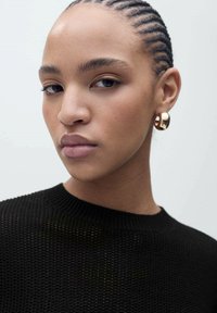 Schwarzer Strickpullover mit rundem Ausschnitt. Nahaufnahme eines Gesichts mit geflochtenem Haar und goldenen Creolen. Glatte Haut und dezentes Make-up.