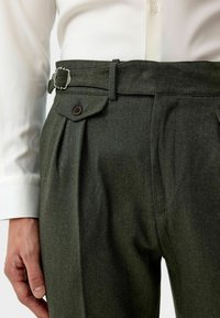 Donkergroene, gestructureerde broeken met een riem met een metalen gesp, voorplooien en een knoopsluiting flapzak. Gecombineerd met een wit shirt.