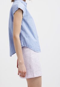 Frau in himmelblauem, kurzärmligem Hemd und strukturiertem, pastell-lila, hoch tailliertem Rock, seitlich vor einem einfarbigen Hintergrund gezeigt.