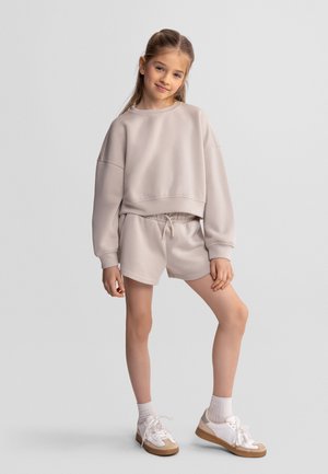 Młoda dziewczyna w beżowej, oversize'owej bluzie i dopasowanych szortach, stojąca z jedną nogą lekko ugiętą na jednolitym tle.