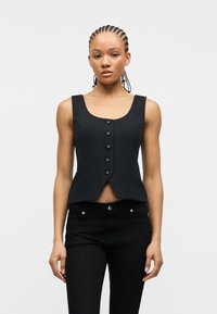 Γυναίκα με πλεξούδες, ντυμένη με αμάνικο μαύρο γιλέκο με κουμπιά και μαύρο παντελόνι, στέκεται μπροστά σε ένα απλό λευκό φόντο.