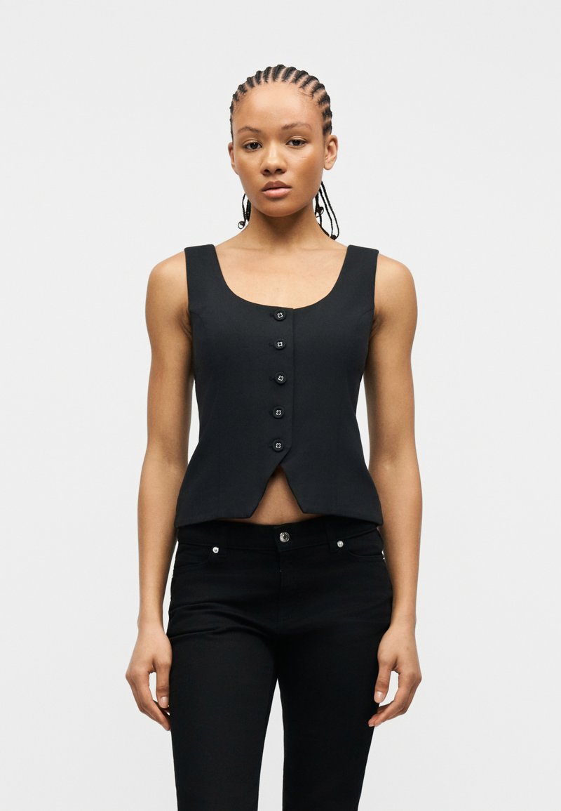 Γυναίκα με πλεξούδες, ντυμένη με αμάνικο μαύρο γιλέκο με κουμπιά και μαύρο παντελόνι, στέκεται μπροστά σε ένα απλό λευκό φόντο.