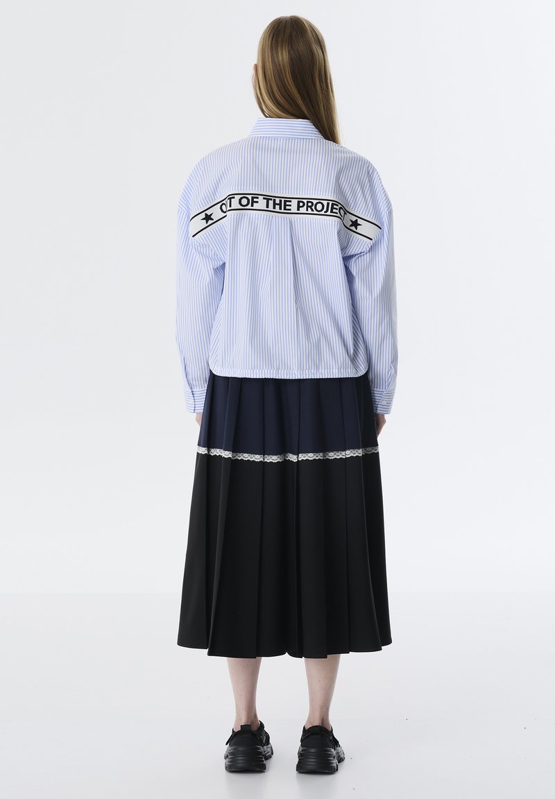 Camisa de botones azul a rayas con texto gráfico negro en la parte posterior, combinada con una falda negra plisada con una banda azul marino y un acento de encaje.