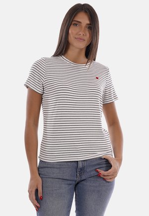 Mujer que lleva una camiseta de manga corta a rayas blancas y negras con un pequeño corazón rojo bordado, combinada con vaqueros azules.