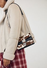 Personne portant une veste en peau lainée beige et une jupe écossaise rouge, portant un sac à motif peau de vache marron et blanc avec une boucle argentée.