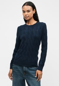 Granatowy sweter dzianinowy z wzorem w warkocze, okrągłym dekoltem i długimi rękawami. ściągacz u dołu i przy mankietach. W zestawie z niebieskimi dżinsami.