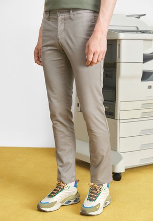 Mann trägt beige Slim-Fit-Hose und bunte Sneaker, steht auf gelbem Teppich neben einem weißen Büro-Drucker.