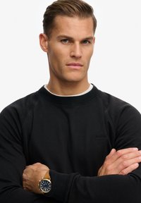 Felpa nera con collo e polsini a costine, caratterizzata da un dettaglio logo sottile. Il modello indossa un orologio d'oro con quadrante blu.