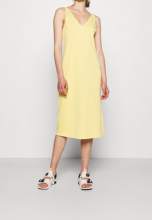Abito giallo senza maniche con scollatura a V, lunghezza al ginocchio, realizzato in tessuto morbido. Indossato con sandali bianchi e neri con più cinturini.