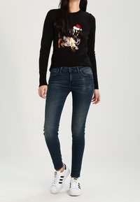 Suéter negro con un gráfico de un perro usando un gorro de Santa, combinado con jeans ajustados de color azul oscuro y zapatillas blancas con rayas negras.