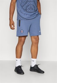Mėlynos Nike šortai su Paryžiaus logotipu, turintys šonines kišenes ir minkštą tekstūrą. Dėvimi su juodais sportiniais batais ir baltais kojiniais.