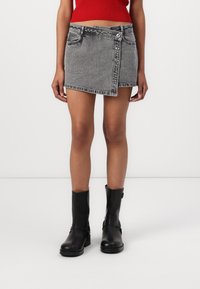Jupe mini en denim gris avec ourlet en angle, fermeture à bouton, deux poches avant, associée à des bottines noires. Texture lisse, design décontracté.