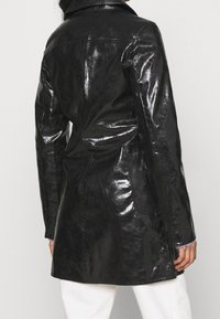 Manteau en cuir noir brillant avec col et taille cintrée. Il comporte des manches longues et des poches latérales, et arrive jusqu'à mi-cuisse.