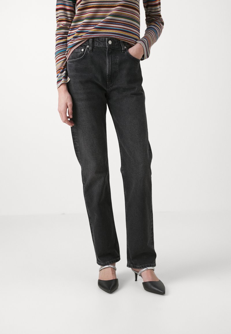 Jeans negros de cintura alta en denim rígido con un corte recto, que cuentan con bolsillos frontales y una sutil textura descolorida. Combinados con tacones negros de punta.