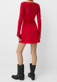 Rød langærmet kjole med tætsiddende liv og flagrende skørt, parret med knæhøje sorte læderstøvler med spænder og struktureret overflade.