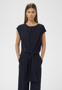 Salopetă de culoare bleumarin, cu mâneci scurte răsucite, guler înalt și talie legată. Fabricată dintr-un material neted și ușor, cu o croială lejeră.