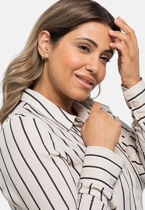 Blusa bianca con righe verticali nere, caratterizzata da un colletto e polsini con bottoni. La modella ha capelli mossi e si sta toccando il viso.