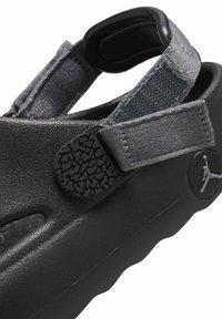 Svart sandal med tjock sula, texturerat syntetmaterial, grå justerbar rem och präglad logodetalj. Har öppna sidor för ventilation.