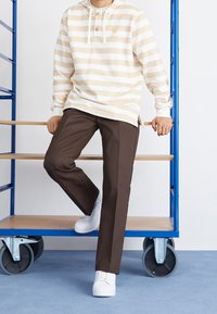 Beige och vit randig hoodie med knappdetalj, kombinerad med bruna byxor och vita sneakers. Visad mot en blå bakgrund.