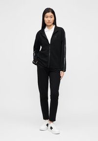 Schwarze Fleecejacke mit Reißverschluss und langen Ärmeln, mit weißem Branding an den Ärmeln. Kombiniert mit schwarzer Hose und weißen Sneaker-Akzenten.