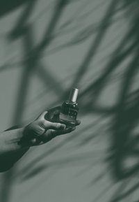 Transparent glass perfume bottle with silver cap held in a hand, featuring a minimalist label and shadowy background with patterned textures.