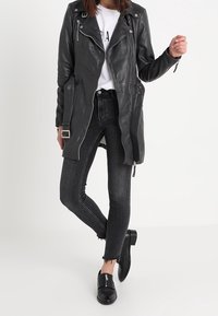 Veste en cuir noir avec fermeture éclair asymétrique, poches latérales et accessoires en métal argenté ; associée à un jean skinny noir et des chaussures slip-on noires.