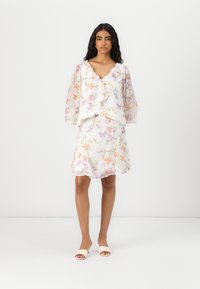 Blouse à imprimé floral avec un col à volants, manches longues translucides, associée à une jupe évasée assortie. Tissu léger et aérien dans des couleurs pastel.