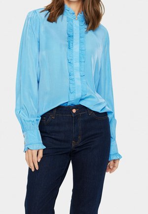 Kvinde iført en lyseblå bluse med lange ærmer og flæsedetaljer samt mørkeblå denimjeans, stående mod en ensfarvet baggrund.