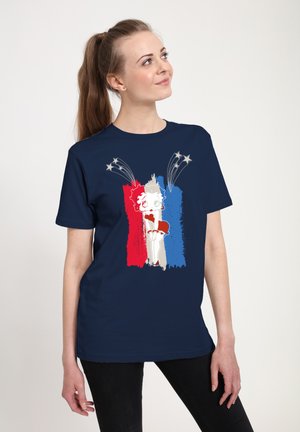 Kobieta z brązowym kucykiem, ubrana w granatową koszulę z wizerunkiem Betty Boop na tle czerwonych, białych i niebieskich pasków oraz lecących gwiazd powyżej.