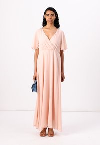 Rochie lungă dintr-un material roz moale, cu decolteu în V, mâneci flutură și talie în stil imperial. Modelul ține în mână o poșetă albastră și poartă sandale bej.