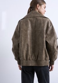 Γυναίκα με ανοιχτό καστανά μαλλιά δεμένα σε αλογοουρά, που φοράει ένα χαλαρό καφέ δερμάτινο μπουφάν με ελαστικό τελείωμα και σκούρο παντελόνι, γυρισμένη πλάτη μπροστά σε λευκό τοίχο.