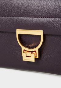 Purple leather handbag featuring a gold metallic handle and clasp, with a textured surface and visible stitching details.