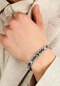 Silverfärgad metallarmband med växlande segment av svarta och blå vävda detaljer, med en central ingraverad emblem.