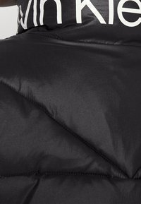 Chaleco acolchado negro con acabado brillante, diseño acolchado y un prominente logo blanco de "Calvin Klein" en el pecho.