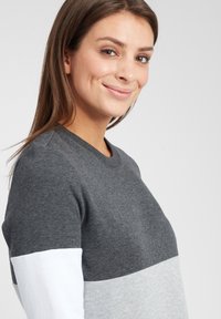 Felpa grigia con maniche lunghe, caratterizzata da un design a blocchi di colore: parte superiore grigia scura, parte media grigia chiara e parte inferiore bianca. Tessuto morbido.