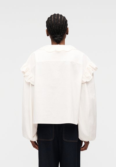 Personne aux cheveux tressés en chignon bas portant un chemisier blanc à manches longues avec des détails volantés aux épaules et un pantalon sombre, vue de dos.