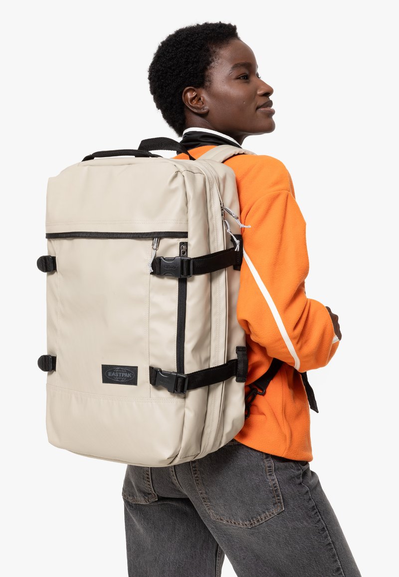 Ljust beige ryggsäck med svarta remmar och detaljer. Rektangulär form, flera zipprade fack och ett handtag på toppen för enkel transport.