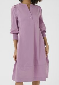 Jasnofioletowa sukienka z bufiastymi rękawami, zapinana na guziki, o luźnym kroju. Wykonana z gładkiego materiału, o długości do kolan.