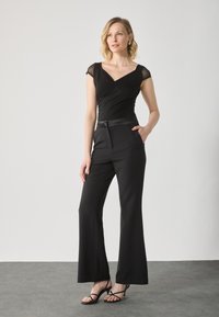 Salopetă neagră cu un top în stil înfășurat, mâneci scurte transparente, talie ajustată cu un brâu din satin și pantaloni largi. Sandale negre cu bretele.